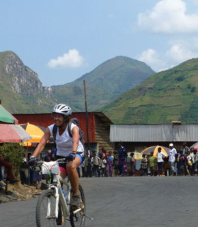 Représentation de l'article, Randonner au Congo, défis des nouveaux aventuriers, une cycliste avec la chaine de volcan dans son dos.